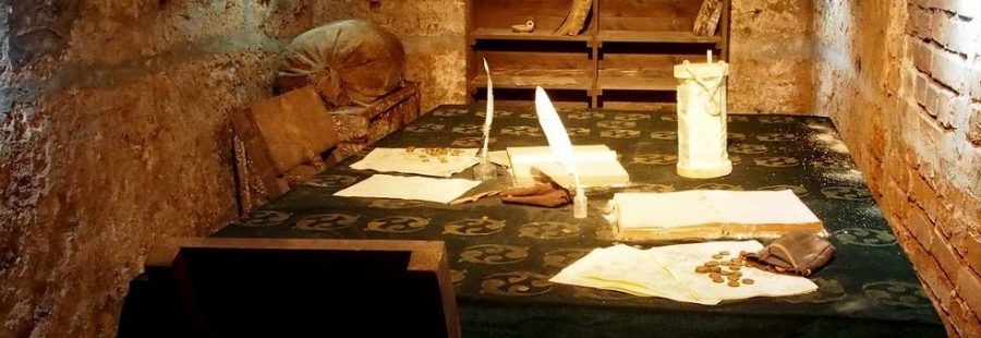 Podziemia w twierdzy Kłodzko, stół z dokumentami i monetami, półki z książkami, worki i krzesła